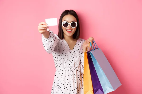 Krásná asijská žena v slunečních brýlích jít nakupovat, držení tašky a ukazuje kreditní kartu, stojící nad růžovým pozadím — Stock fotografie