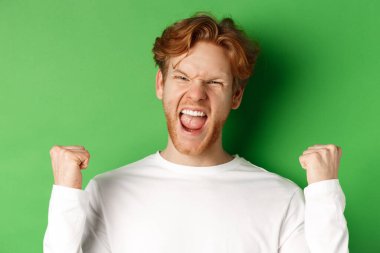 Şanslı genç adamın yakın çekimi, zaferi kutluyor, sevinçle evet diye bağırıyor ve yumruk sallıyor, yeşil arka planda zafer kazanıyor.