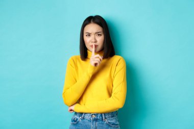 Hayal kırıklığına uğramış Asyalı bir kadın, sessiz olmasını söylüyor, yüksek sesle bağıran birini susturuyor, kameraya şşşş yapıyor, üzgün üzgün bakıyor, mavi arka planda dikiliyor.
