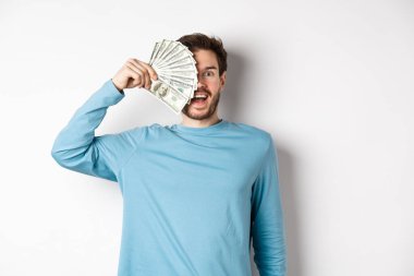 Şaşırmış ve mutlu genç adam yüzünün yarısını parayla kaplıyor, kameraya hayret ediyor, hızlı kredi tekliflerini kontrol ediyor, beyaz arka planda duruyor.