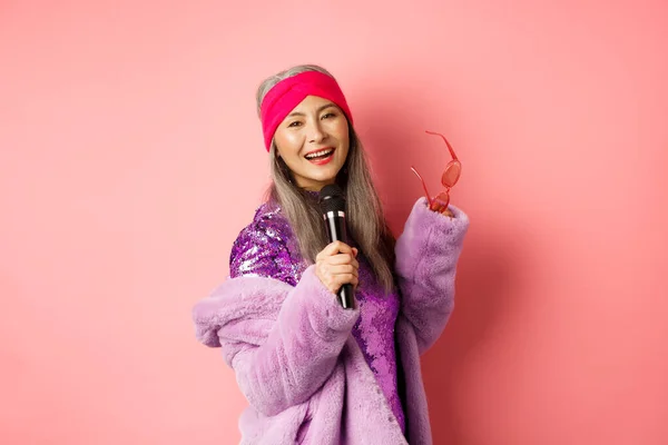 Linda asiática sênior mulher cantando karaoke no microfone, executar música e olhando feliz, de pé sobre fundo rosa — Fotografia de Stock