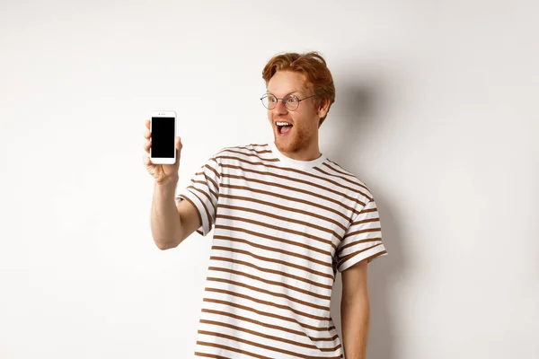 Concepto de tecnología y comercio electrónico. Feliz joven pelirrojo en gafas que muestran la pantalla del teléfono inteligente en blanco, mirando sorprendido, de pie sobre fondo blanco — Foto de Stock