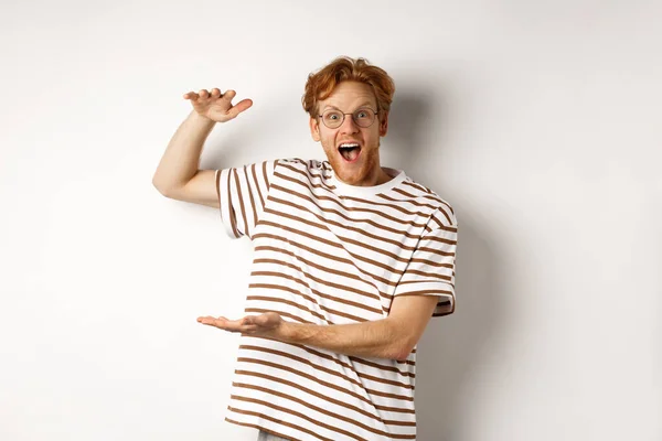 Amazed redhead man in glasses showing something large size and smiling at camera, saying wow, standing over white background — Stock Photo, Image