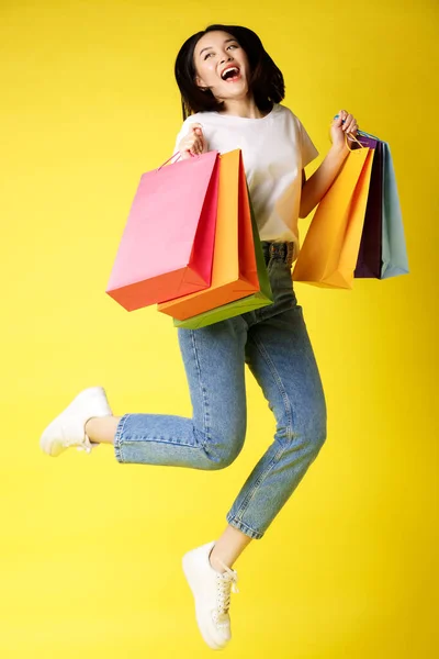 Full size shot der schönen asiatischen Frau springt mit Einkaufstüten und lächelt glücklich, gelber Hintergrund — Stockfoto