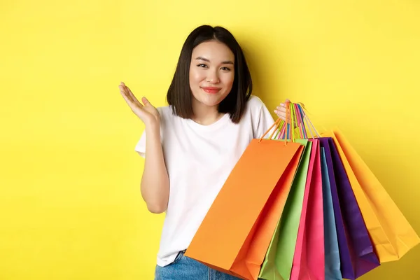 Hloupé asijské dívka ukazuje nákupní tašky a pokrčení rameny, obchod v obchodech se slevami, stojící nad žlutým pozadím — Stock fotografie