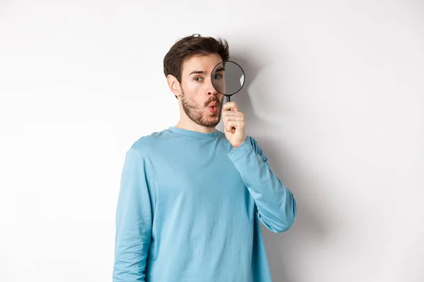 Surprised man found something, looking through magnifying glass with amazed face, standing over white background — Stock Photo, Image