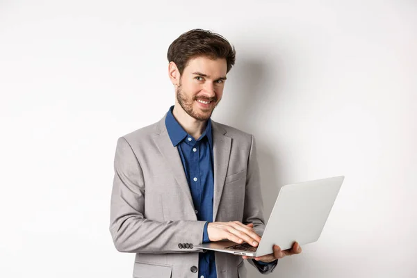 Succesvolle lachende zakenman werkend op laptop en kijkend blij op camera, staand in grijs pak op witte achtergrond — Stockfoto