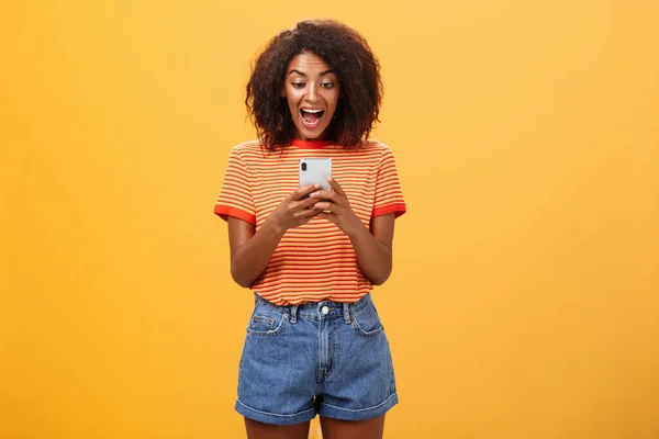 Chica expresando emoción y alegría recibiendo una invitación impresionante a través de mensajes gritando desde el placer y la felicidad mirando la pantalla del teléfono inteligente impresionado y emocionado sobre el fondo naranja — Foto de Stock