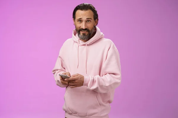 Stylische gut aussehende erwachsene bärtige Mann 40er Jahre in trendigen rosa Kapuzenpullover mit Smartphone-SMS-Look Kamera unbehelligt cool selbstbewusst abgelenkt spielen awesome neues App-Spiel, lila Hintergrund — Stockfoto