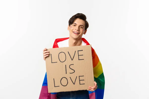 Smiling gay man activist holding sign love is love for lgbt pride parade, wearing Rainbow flag, standing over white background — Stock Photo, Image