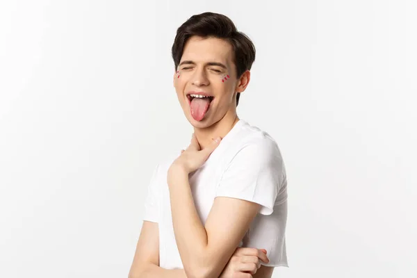 People, lgbtq and beauty concept. Close-up of carefree gay man with glitter on face, showing tongue and making funny face, standing over white background — Stock Photo, Image