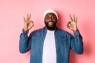 Hipster bereli tatmin olmuş Afro-Amerikan bir adam, onaylama işaretleri gösteriyor, iyi bir teklifi övüyor ve evet diyor, mutlu gülümsüyor, pembe arka planda duruyor.