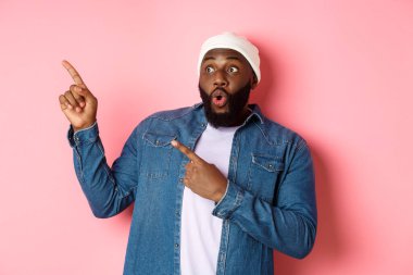 Şapkalı ve kot gömlekli etkileyici hippi siyahi adam sol üst köşedeki logoya bakıyor ve işaret ediyor, şaşkın şaşkın bakıyor, pembe arka planda duruyor.