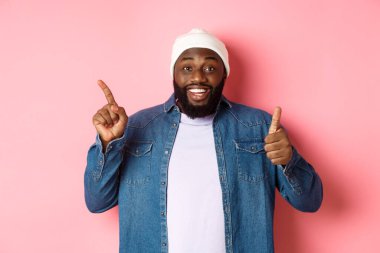 Mutlu Afro-Amerikalı adam baş parmağını yukarı kaldırıyor ve sol üst köşedeki fotokopi alanını işaret ediyor, iyi bir anlaşmaya övgüler yağdırıyor, pembe arka planda duruyor.