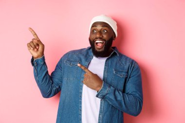 Şaşırmış siyahi adam harika bir reklam sunuyor, gülümsüyor ve parmaklarını sol üst köşedeki pankarta gösteriyor, pembe arka plan.