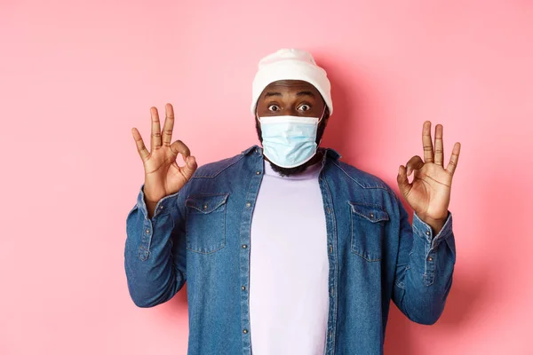Coronavirus, τρόπος ζωής και την κοινωνική έννοια αποστασιοποίηση. Εντυπωσιασμένος Αφροαμερικάνος με μάσκα προσώπου, που δείχνει εντάξει σημάδι, εγγύηση και έπαινο, στέκεται πάνω από ροζ φόντο — Φωτογραφία Αρχείου