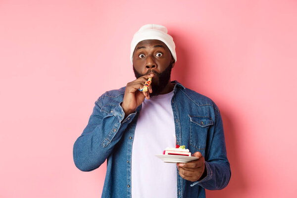 Happy Black hipster celebrating birthday, blowing party whistle, holding bday cake with candle, standing over pink background