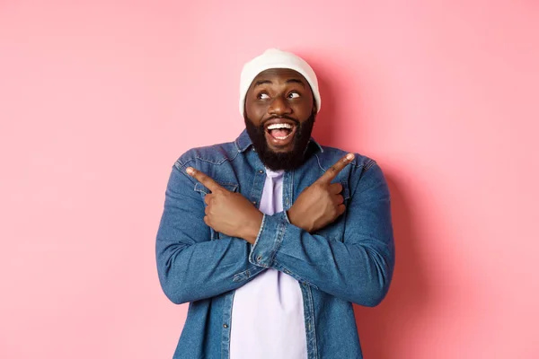 Aufgeregter junger afrikanisch-amerikanischer Mann zeigt zur Seite, zeigt zwei Entscheidungen und trifft Entscheidungen, lächelt glücklich, steht über rosa Hintergrund — Stockfoto