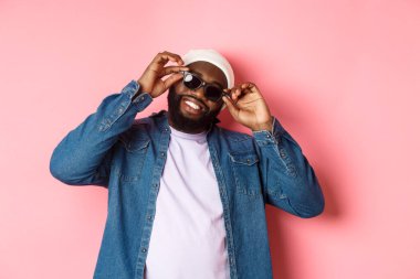 Yakışıklı Afro-Amerikalı adam güneş gözlüğü takmış, gülümsüyor memnun ve kendinden emin, pembe arka plana karşı duruyor.