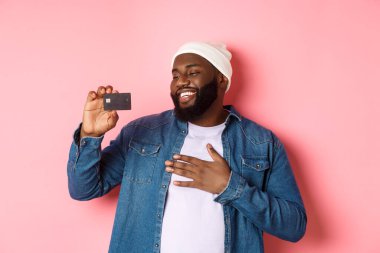 Alışveriş konsepti. Mutlu Afro-Amerikalı bir adamın resmi kredi kartından memnun görünüyor, bankayı tavsiye ediyor, pembe arka planda duruyor.