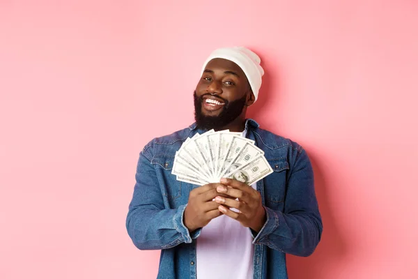 Satisfait homme riche afro-américain montrant de l'argent, souriant heureux et frimeur avec des revenus, debout avec des dollars sur fond rose — Photo