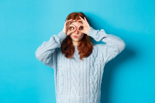 Süveter giyen komik kızıl saçlı kadın model, kameraya parmak gözlükleriyle bakıyor, mavi arka planda dikilen ilgi ve şaşkınlığını dile getiriyor. — Stok fotoğraf