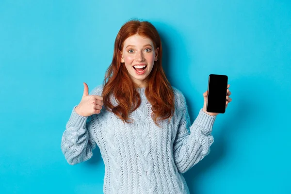Улыбающаяся рыжая девушка показывает экран смартфона, держит телефон и демонстрирует приложение, делает большой палец вверх в утверждении, рекомендуя интернет-магазин, синий фон — стоковое фото