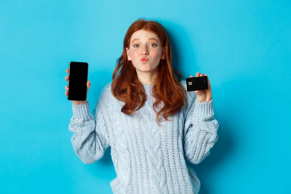 兴奋的红头发女孩站在蓝色背景上，展示手机屏幕和信用卡，演示网上商店或应用程序 — 图库照片