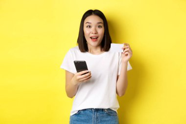 E-ticaret ve online alışveriş konsepti. Heyecanlı Asyalı kadın internetten sipariş veriyor, elinde akıllı telefon ve plastik kredi kartı var, sarı arka planda duruyor.