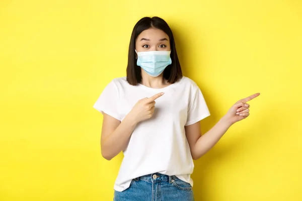 Covid-19, pandemiskt och socialt distanserande koncept. Förvånad och glad asiatisk kvinna i medicinsk mask, visar reklam, pekar höger och leende, gul bakgrund — Stockfoto