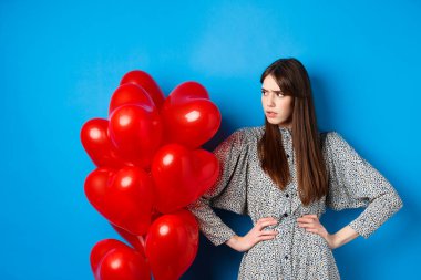 Sevgililer Günü. Sinirli ve rahatsız bir kız şikayetçi, boş alana bakıyor ve somurtuyor, kalp balonlarının yanında duruyor, mavi arka plan