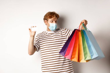 Covid, karantina ve yaşam tarzı konsepti. Bankanın plastik kredi kartıyla alışveriş torbalarına bakan kızıl saçlı hayret verici bir adam.