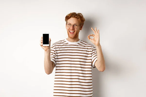 Concepto de tecnología y comercio electrónico. Hombre joven con el pelo rojo mostrando signo de bien y pantalla de teléfono inteligente en blanco, alabando aplicación impresionante, de pie sobre fondo blanco — Foto de Stock
