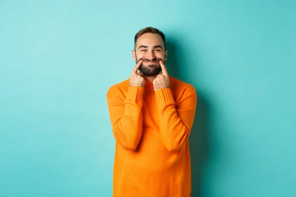 Image of bearded man stretching lips in happy smile, faking happiness, standing over light blue background — Stock Photo, Image