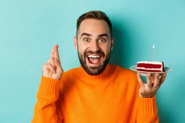 Close-up of happy adult man celebrating birthday, holding bday cake with candle and making wish, standing against turquoise background — Stock Photo, Image