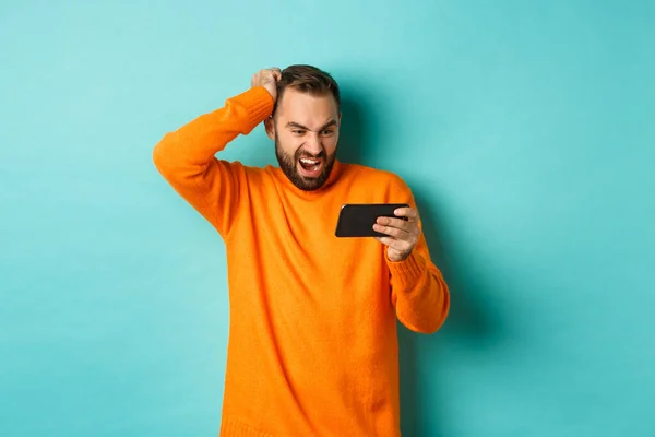 Immagine di uomo guardando confuso al telefono cellulare, guardando qualcosa online, in piedi perplesso sullo sfondo azzurro — Foto Stock