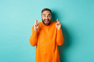 Heyecanlı adam beklenen bir şey bekliyor, parmaklarını çapraz yapıp gülümsüyor, dilek tutuyor, açık mavi arka planda duruyor.