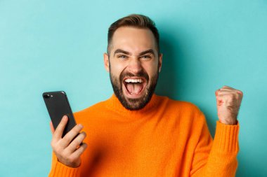 Cep telefonuyla kazanan adamın görüntüsü, yumruk tokuşturma ve zaferin sevinci, akıllı telefonu tatmin olmuş bir yüzle tutmak, turkuaz arka plan