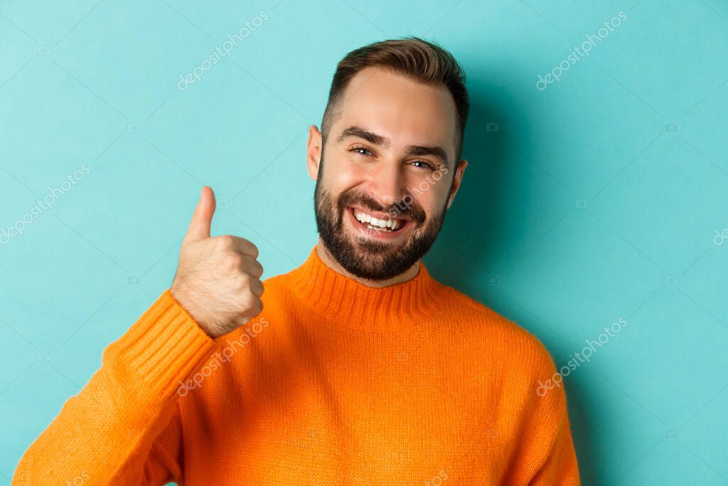 Close-up of attractive young man showing thumb-up, praising good work, standing over light blue background, giving positive answer.