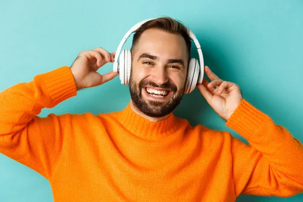 Kulaklıkla müzik dinleyen, turuncu süveter giymiş turkuaz arka planda duran yakışıklı modern adamın yakın çekimi. — Stok fotoğraf