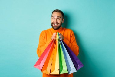 Turuncu kazaklı mutlu beyaz adam, sola bakıyor ve görüntülüyor, alışveriş torbaları tutuyor ve gülümsüyor, turkuaz arka planda dikiliyor.