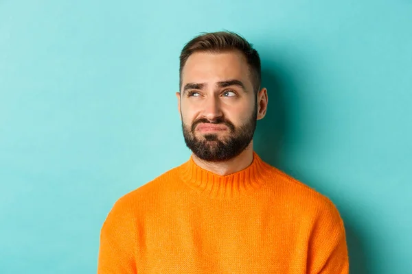 Close-up de belo homem caucasiano olhando para a esquerda decepcionado e cético, olhando para o logotipo, vestindo suéter laranja, de pé contra fundo turquesa — Fotografia de Stock