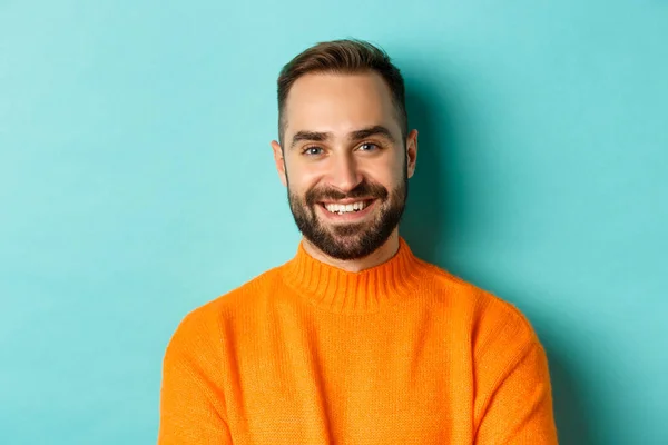 Close-up de belo homem caucasiano sorrindo para a câmera, olhando confiante, vestindo suéter laranja, de pé contra fundo turquesa — Fotografia de Stock