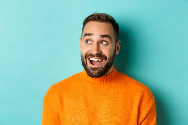 Close-up de belo homem caucasiano sorrindo, olhando para a esquerda com o rosto surpreso, olhando para o logotipo, vestindo suéter laranja, de pé contra fundo turquesa — Fotografia de Stock