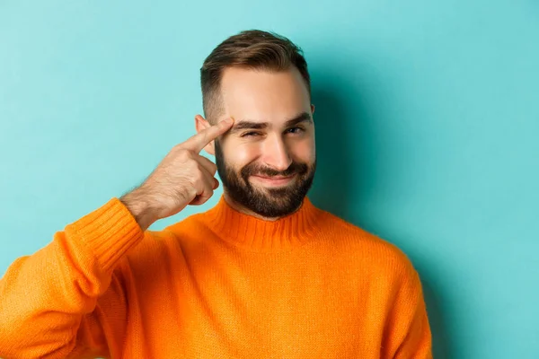 Imagem do jovem em camisola laranja, sorrindo e apontando para a cabeça, tendo uma ideia, elogiando o bom pensamento, de pé sobre fundo azul claro — Fotografia de Stock