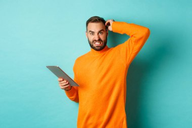 Kafası karışmış, endişeli görünen, dijital tablet üzerinde çalışan, turkuaz arka plana karşı duran bir adam.