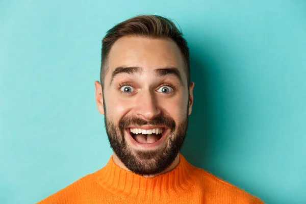 Headshot de belo homem caucasiano com barba sorrindo feliz para a câmera, olhando espantado, de pé em suéter laranja contra fundo turquesa — Fotografia de Stock