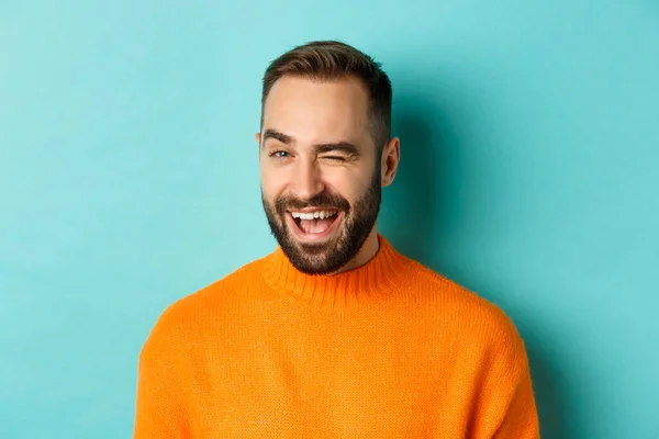 Bonito homem confiante piscando para a câmera, sorrindo atrevida, de pé em suéter laranja contra fundo azul — Fotografia de Stock