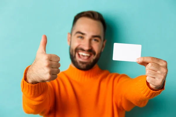 Close-up van knappe blanke man die gaat winkelen, met creditcard en duim omhoog in goedkeuring, staande over turquoise achtergrond — Stockfoto