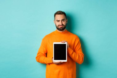 Teknoloji. Şüpheci ve hoşnutsuz adam dijital tablet ekranı gösteriyor, hayal kırıklığına uğruyor, açık mavi arka planda duruyor.